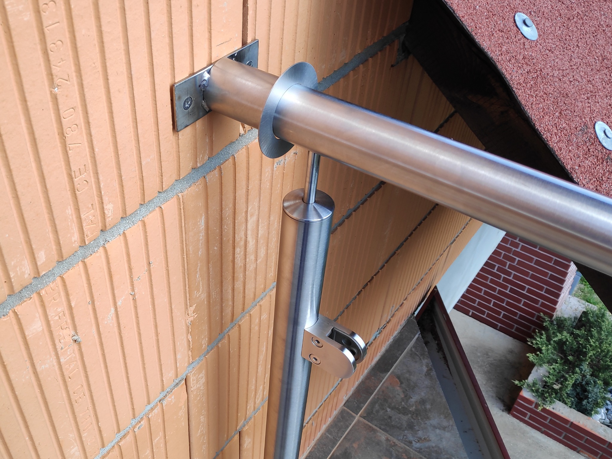 Detale montażu zewnętrznej poręczy ze stali nierdzewnej, mocowanej do ściany z cegły ceramicznej, widoczne elementy mocujące i fragment dachu.