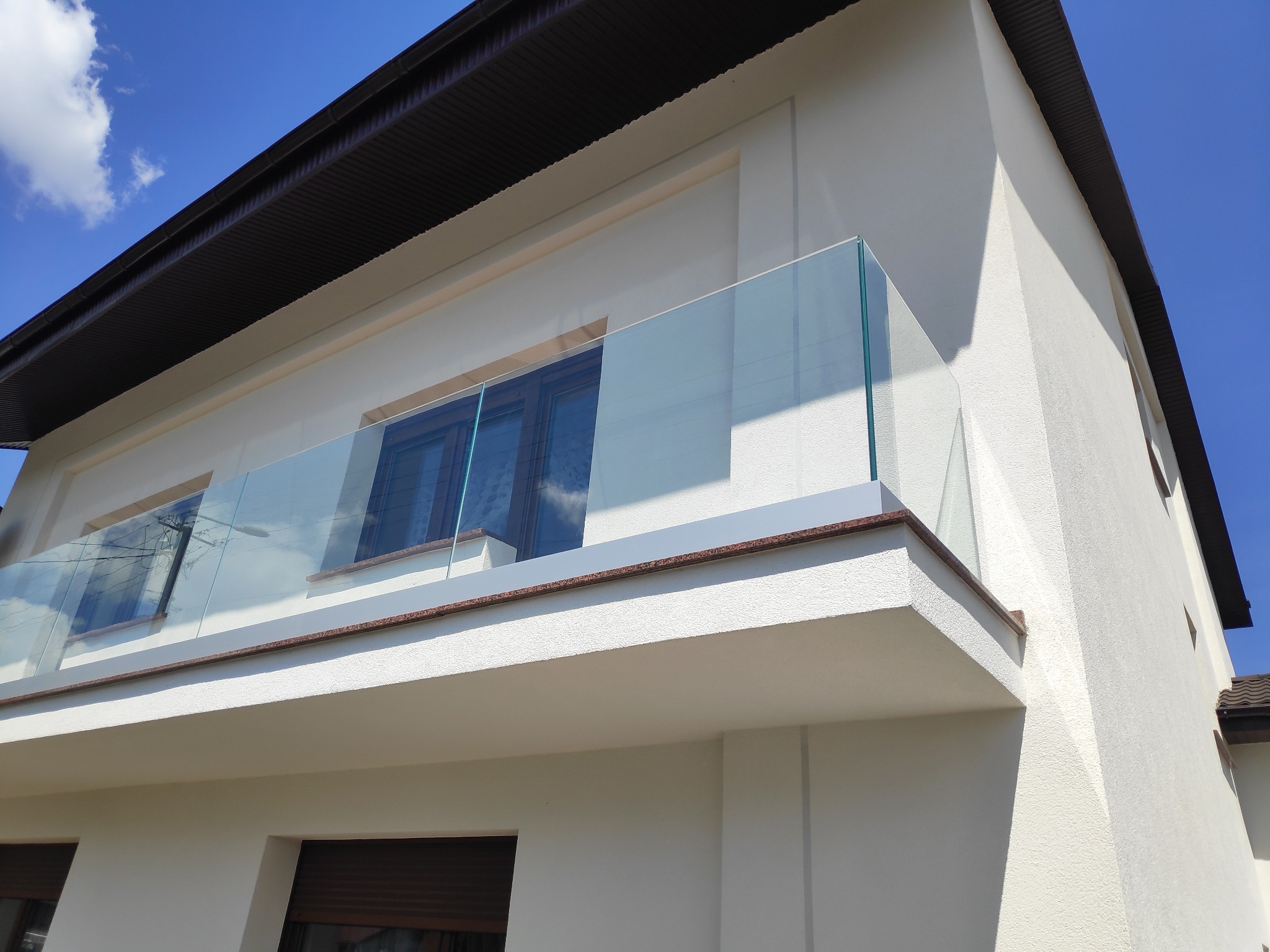 Nowoczesny balkon z przezroczystą taflą szkła, zabezpieczającą przestrzeń, z widocznym odbiciem nieba i instalacji elektrycznej w szybie, na tle jasnej elewacji budynku i błękitnego nieba.