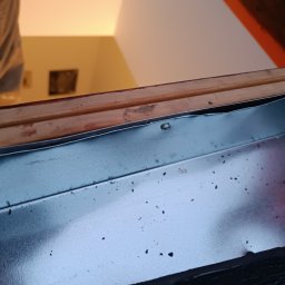 Jacek Dziubaty - Metalowy okapnik okna dachowego przymocowany do drewnianej ramy, widoczne nity mocujące.