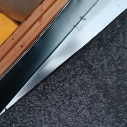 Jacek Dziubaty - Fragment dachu z papą, drewnianą konstrukcją i metalowym obrzeżem, widoczne łączenie materiałów pod kątem.