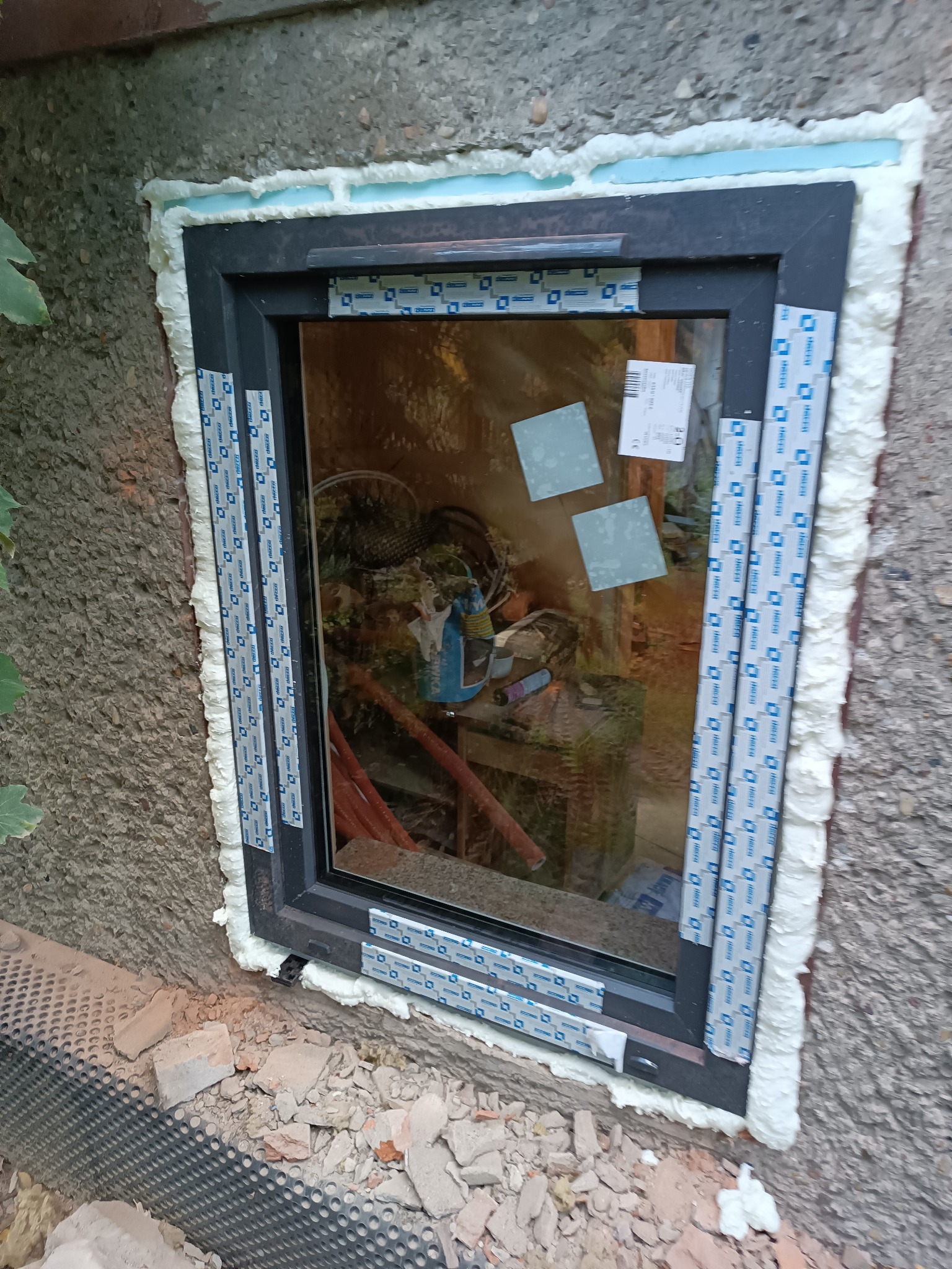 Nowo osadzone okno w szarej ramie z widoczną pianką montażową i taśmą zabezpieczającą, osadzone w surowej ścianie z widocznymi resztkami gruzu.
