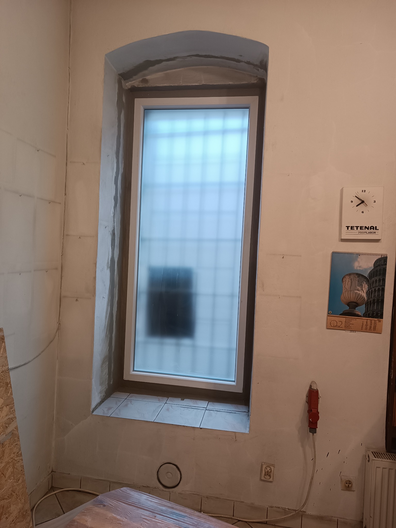 Montaż okna z zaokrąglonym łukiem w surowym stanie, widoczne ślady zaprawy, kalendarz i zegar na ścianie, wystająca wtyczka elektryczna.