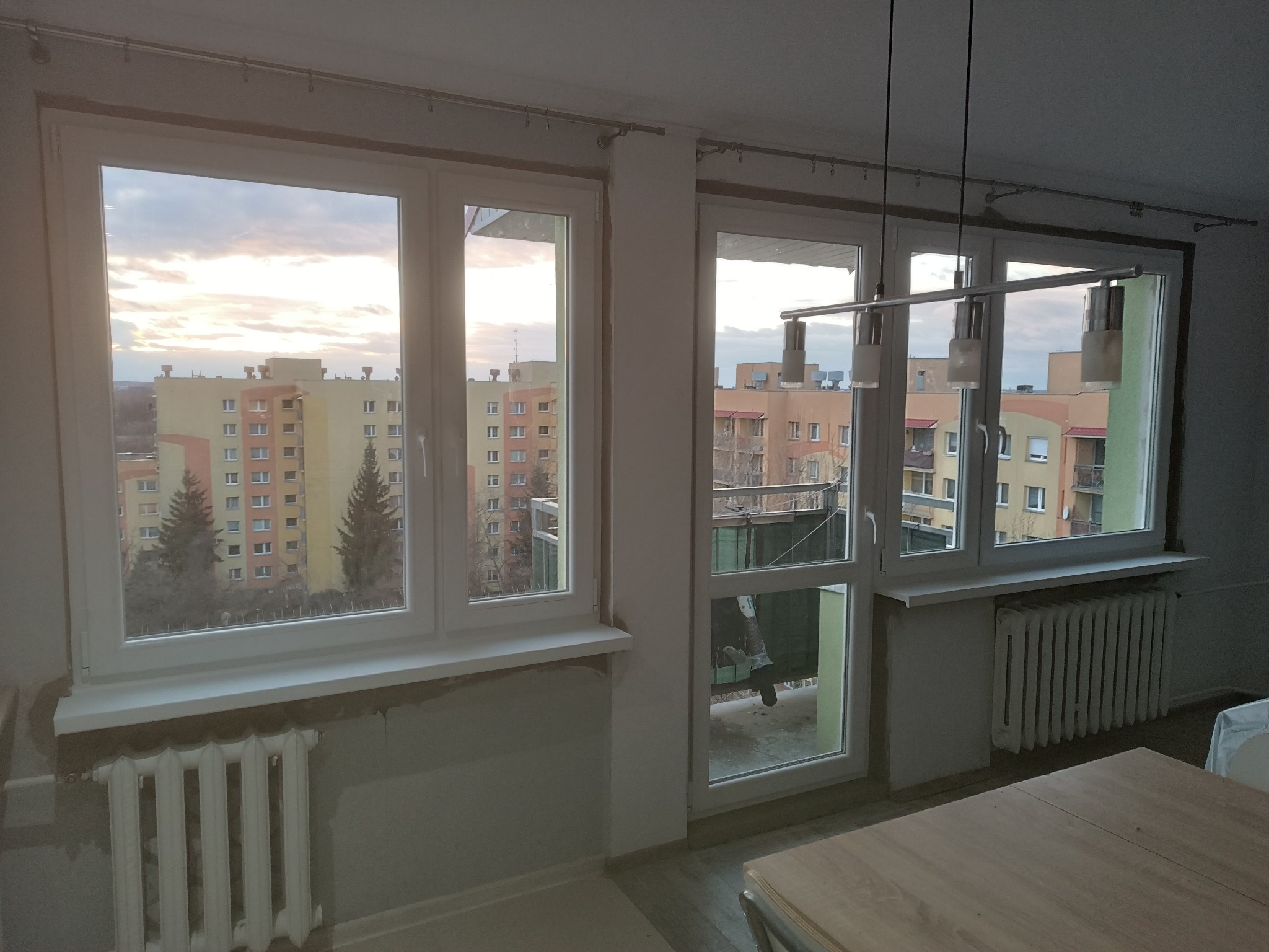 Wnętrze pokoju z dwoma oknami i wyjściem na balkon, widoczne grzejniki i lampa sufitowa, ściany w trakcie prac wykończeniowych.