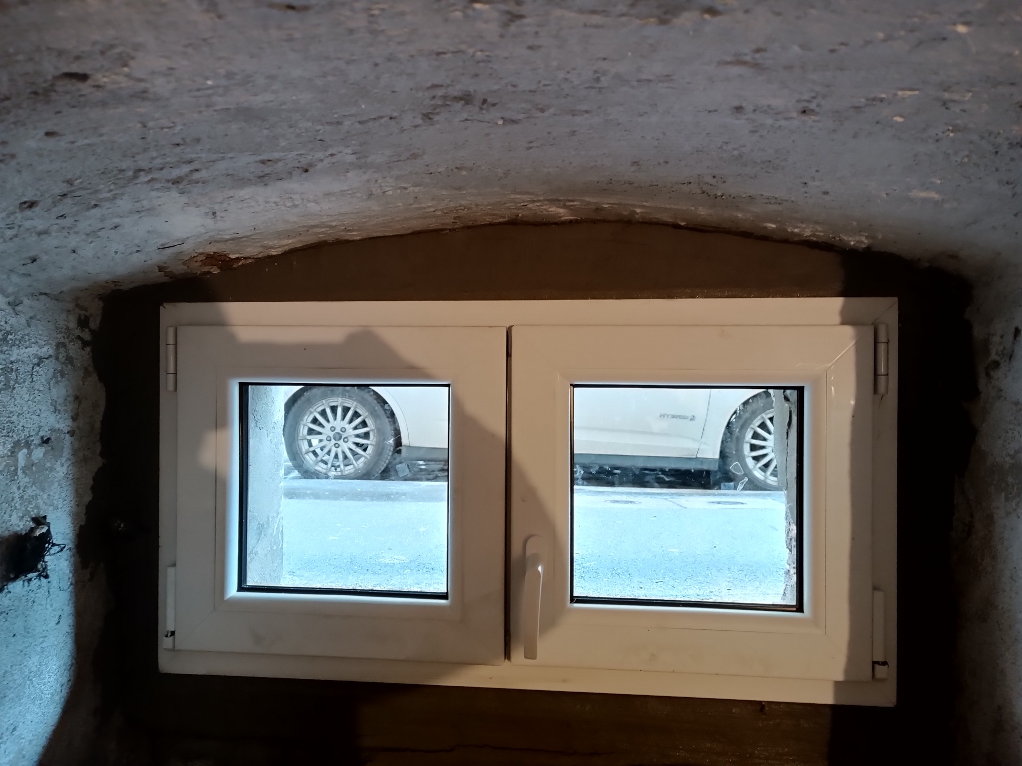 Nowe białe okno PCV z podwójnymi szybami, zamontowane w surowej, szarej ścianie piwnicy, widok na fragment zaparkowanego samochodu na zewnątrz.