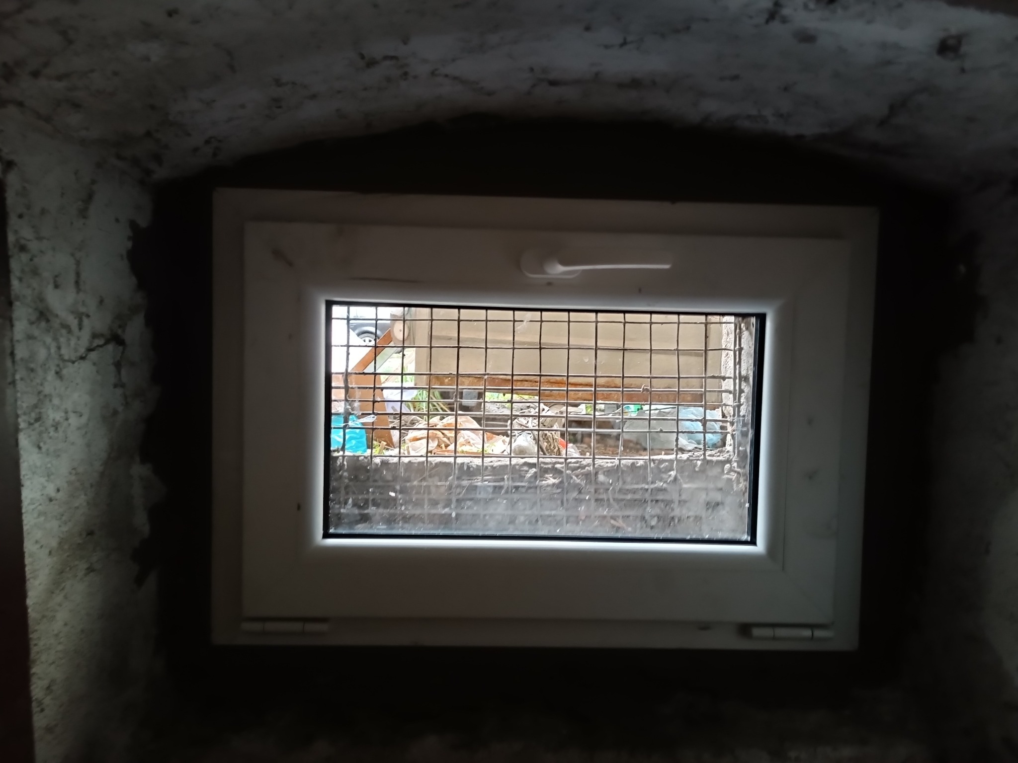 Nowe białe okno z kratą, zamontowane w surowej, betonowej wnęce piwnicznej, widok z wnętrza na zewnątrz, gdzie widać śmieci i gruz.