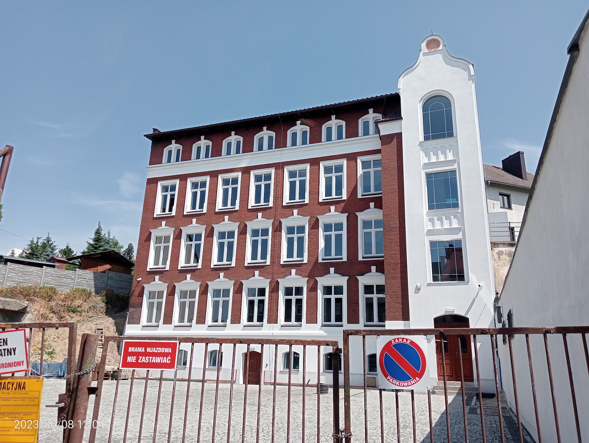 Odnowiona elewacja budynku z czerwonej cegły z białymi detalami, widoczna przez metalową bramę z tabliczką 'Zakaz Parkowania'.