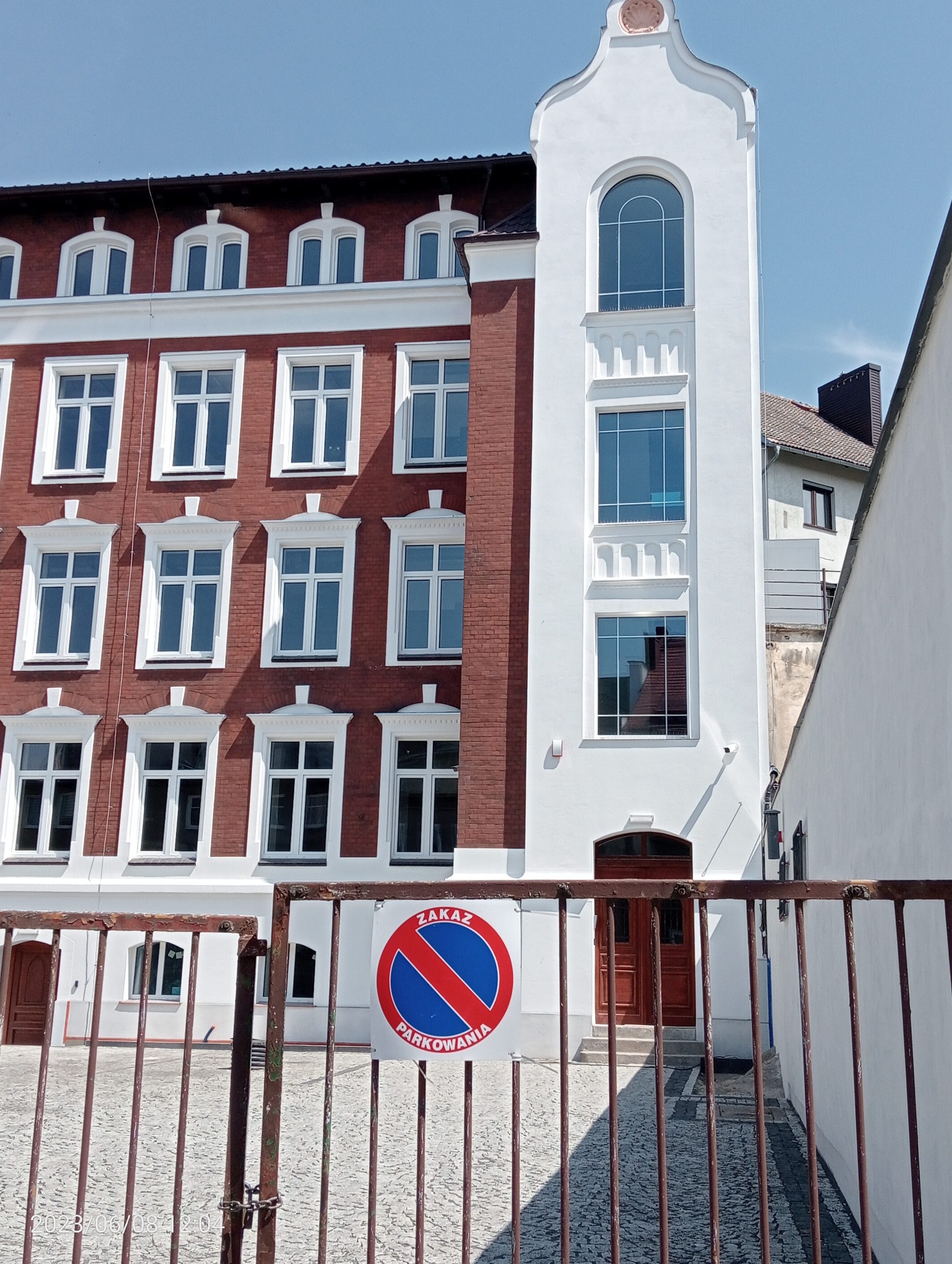 Odnowiona fasada budynku z czerwonej cegły i białego tynku, widoczna przez metalową bramę z tablicą 'Zakaz Parkowania'.