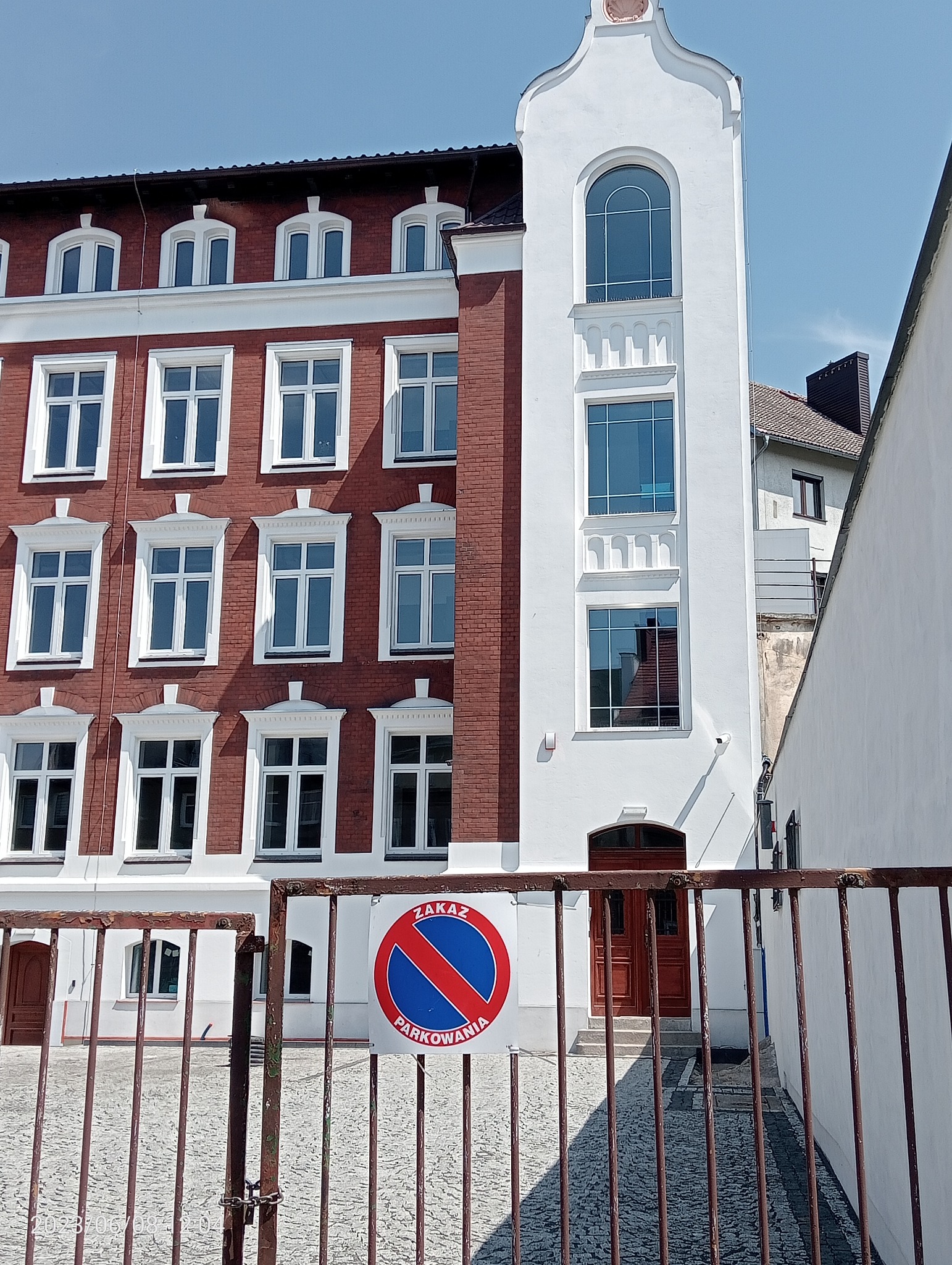 Odnowiony budynek z czerwonej cegły i białego tynku, widoczny przez metalowe ogrodzenie z tabliczką zakazu parkowania.