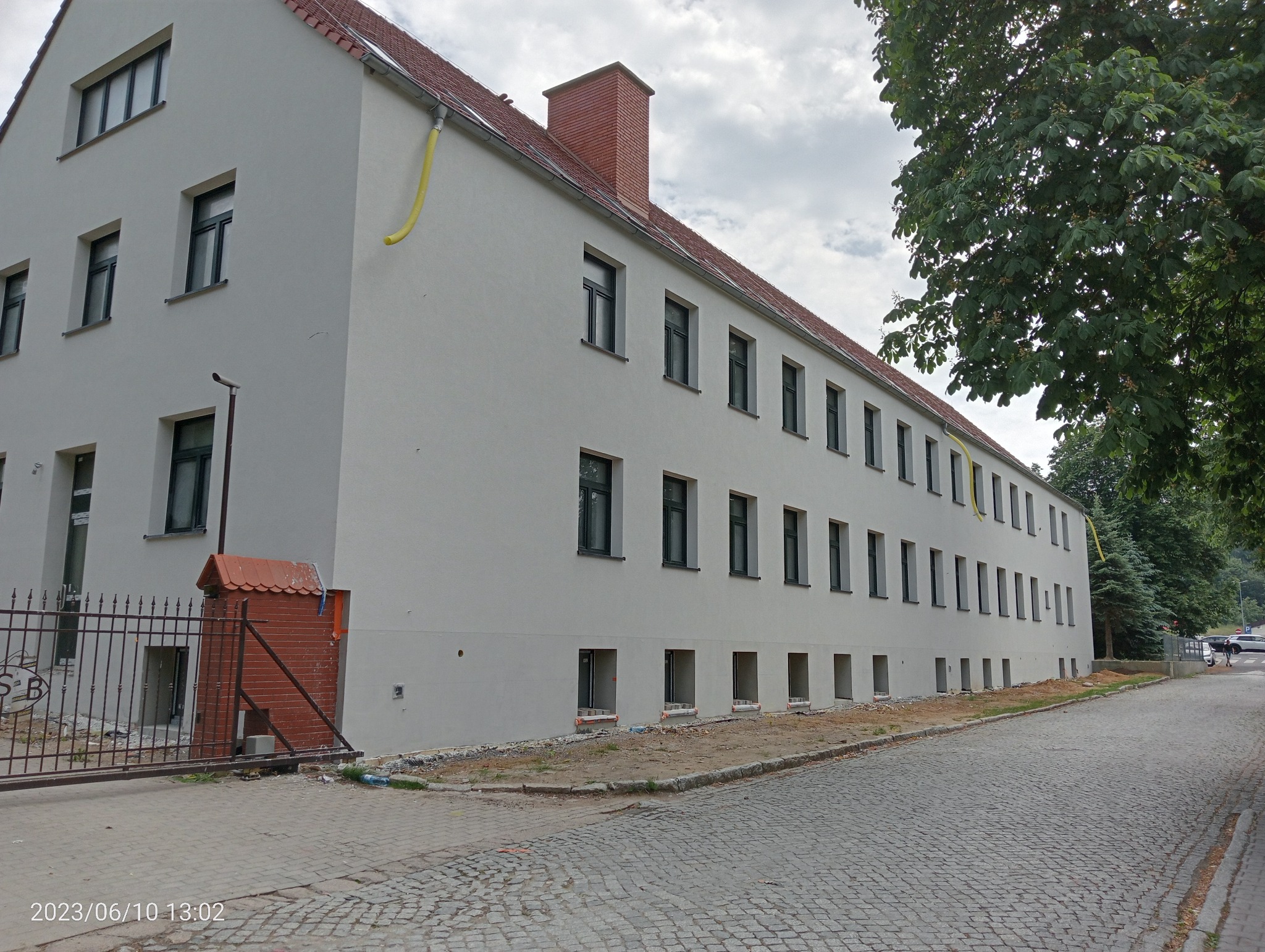 Długi, dwupiętrowy budynek z jasną elewacją i rzędem okien, widoczny z boku, z żółtymi rurami spustowymi wzdłuż dachu, ceglany komin, brukowana ulica i drzewa w tle.