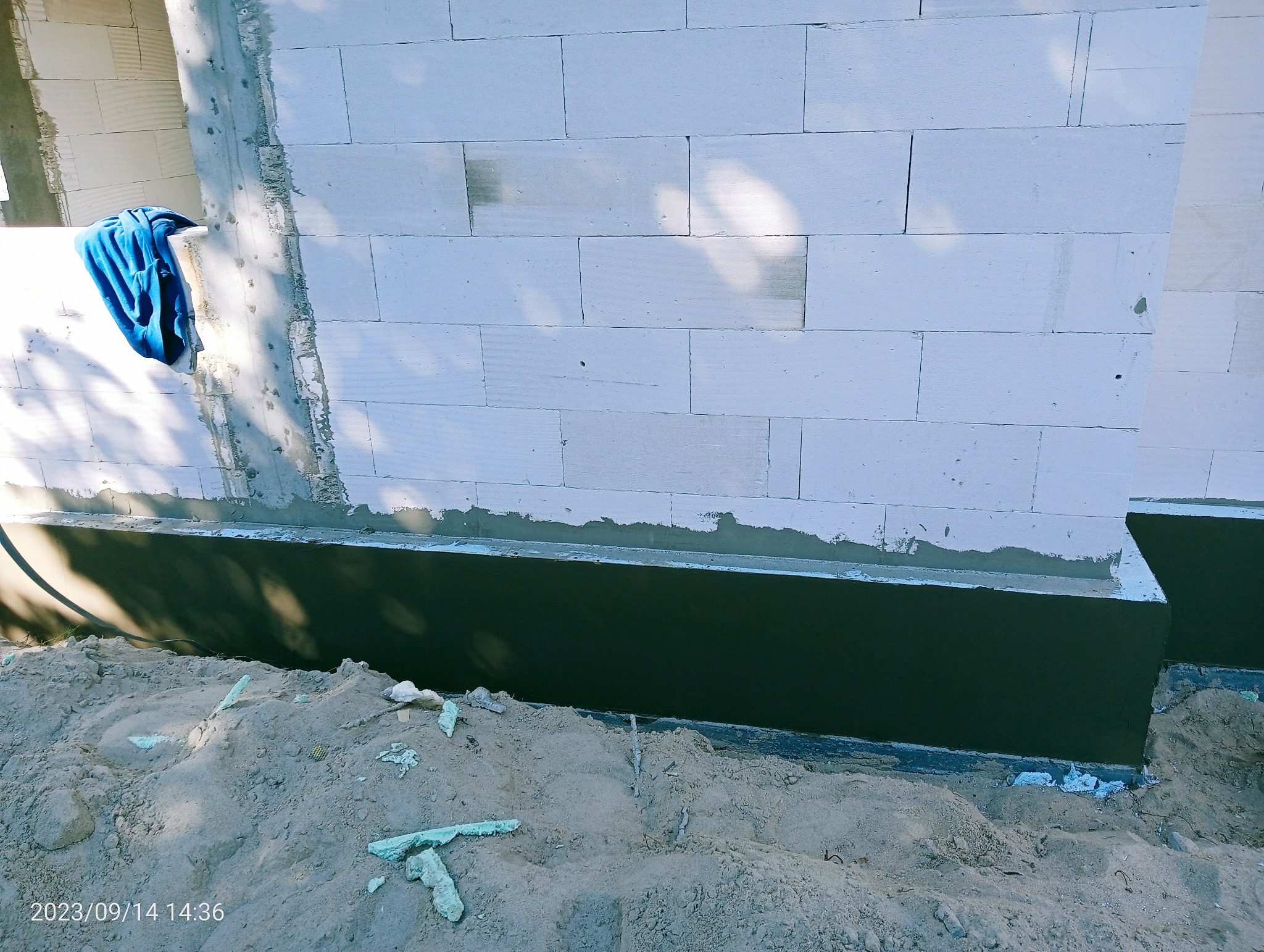 Fundament budynku w trakcie izolacji, widoczna świeża warstwa izolacyjna na fundamencie, obok sterta piasku i fragmenty styropianu, na ścianie budynku leży niebieska szmatka.