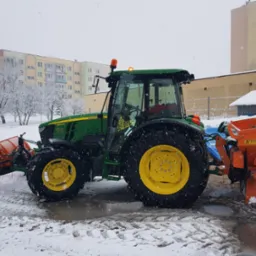 Zielony traktor John Deere z pomarańczowym pługiem i posypywarką stoi na ośnieżonym placu w miejskiej scenerii, podczas opadów śniegu.