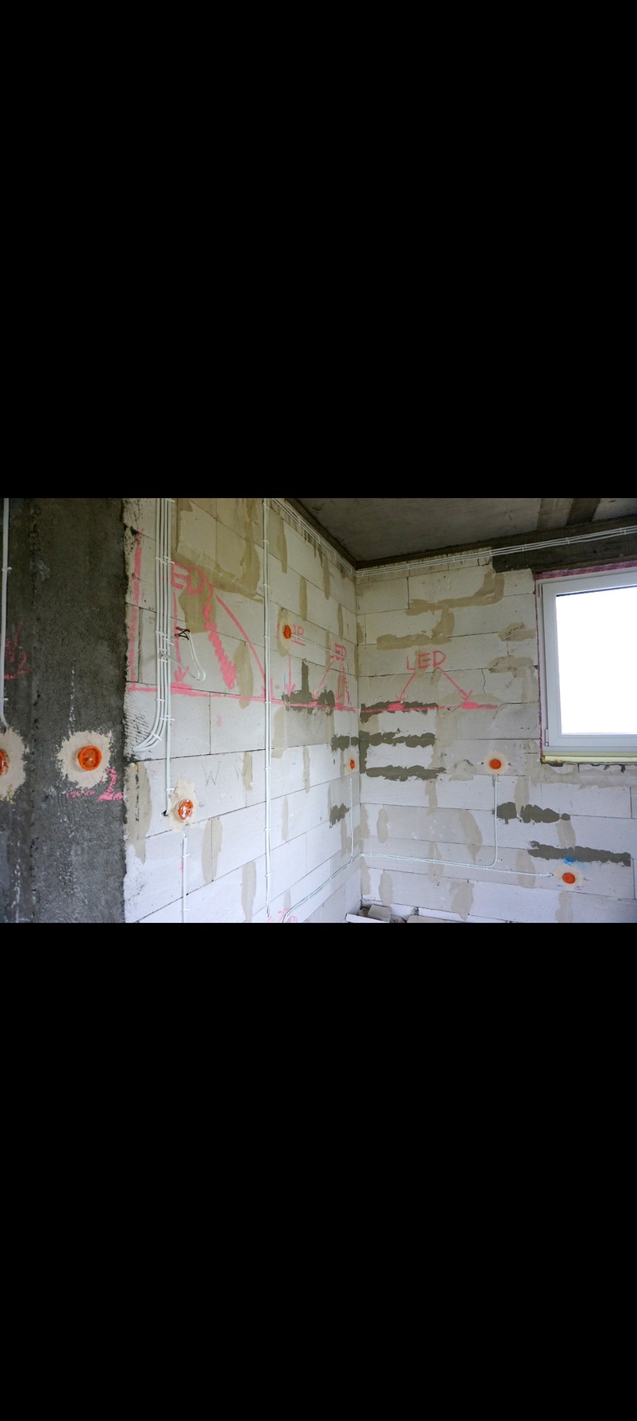 Wnętrze w surowym stanie z widocznymi przewodami elektrycznymi poprowadzonymi po ścianach z betonu komórkowego, zaznaczone miejsca pod gniazdka i oświetlenie LED, różowe oznaczenia na ścianach.