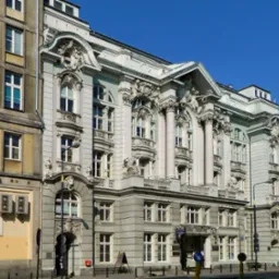 Fasada budynku biurowego z elementami architektonicznymi, kolumnami i zdobieniami, sąsiadująca z budynkiem banku.