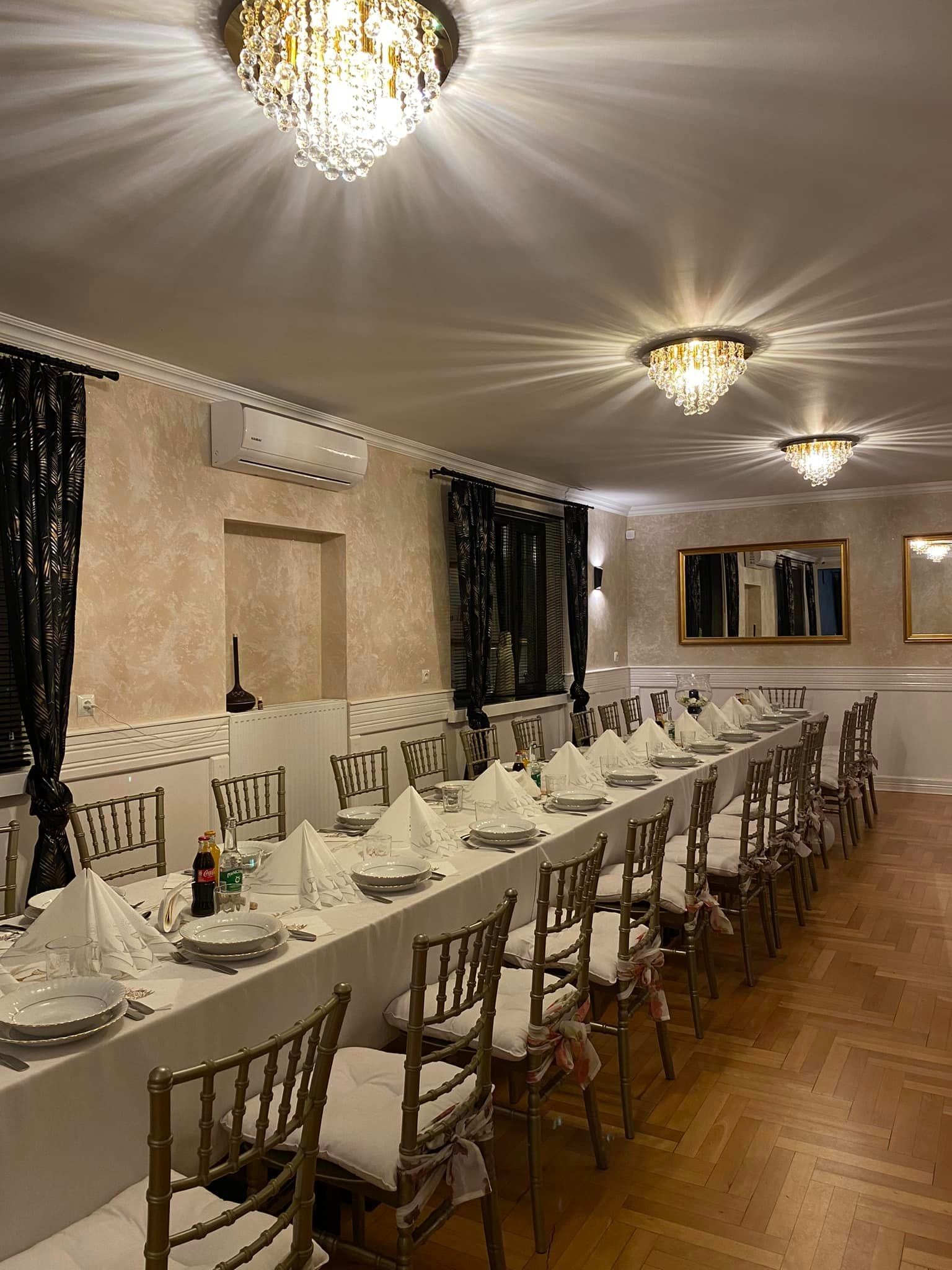 Uroczysta sala z długim stołem nakrytym białym obrusem, zastawą, napojami, i krzesłami z dekoracyjnymi wstążkami, oświetlona kryształowymi żyrandolami.