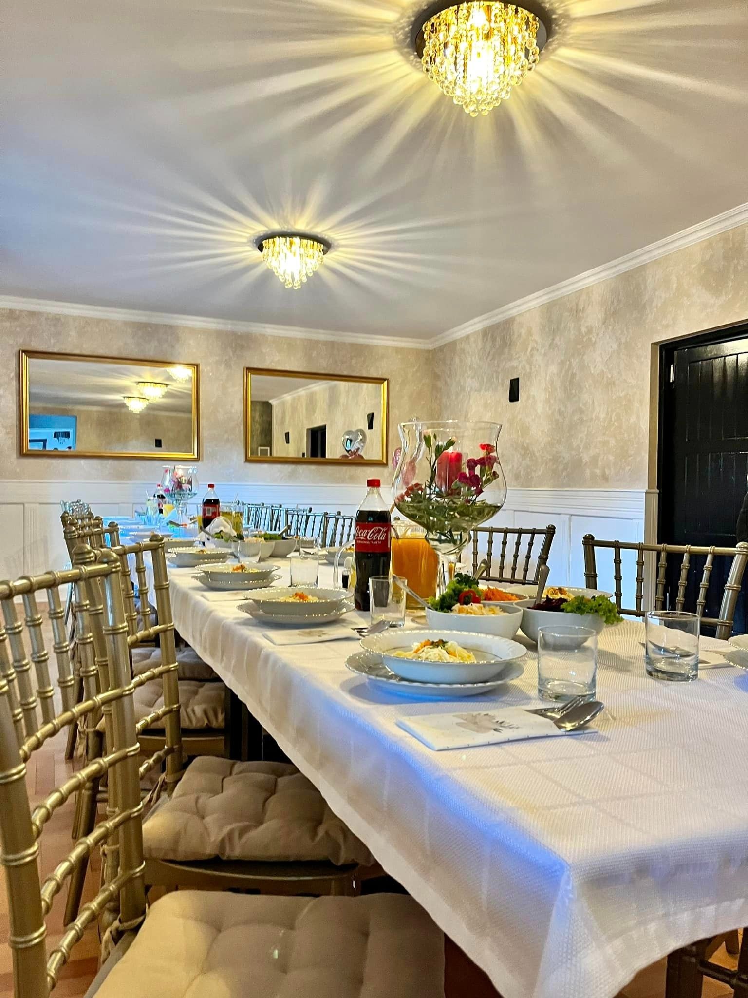 Długi stół nakryty białym obrusem, zastawiony talerzami z daniami, szklankami, napojami w butelkach i dzbankach, udekorowany kwiatami w szklanym wazonie. Krzesła w kolorze złotym ustawione wzdłuż...