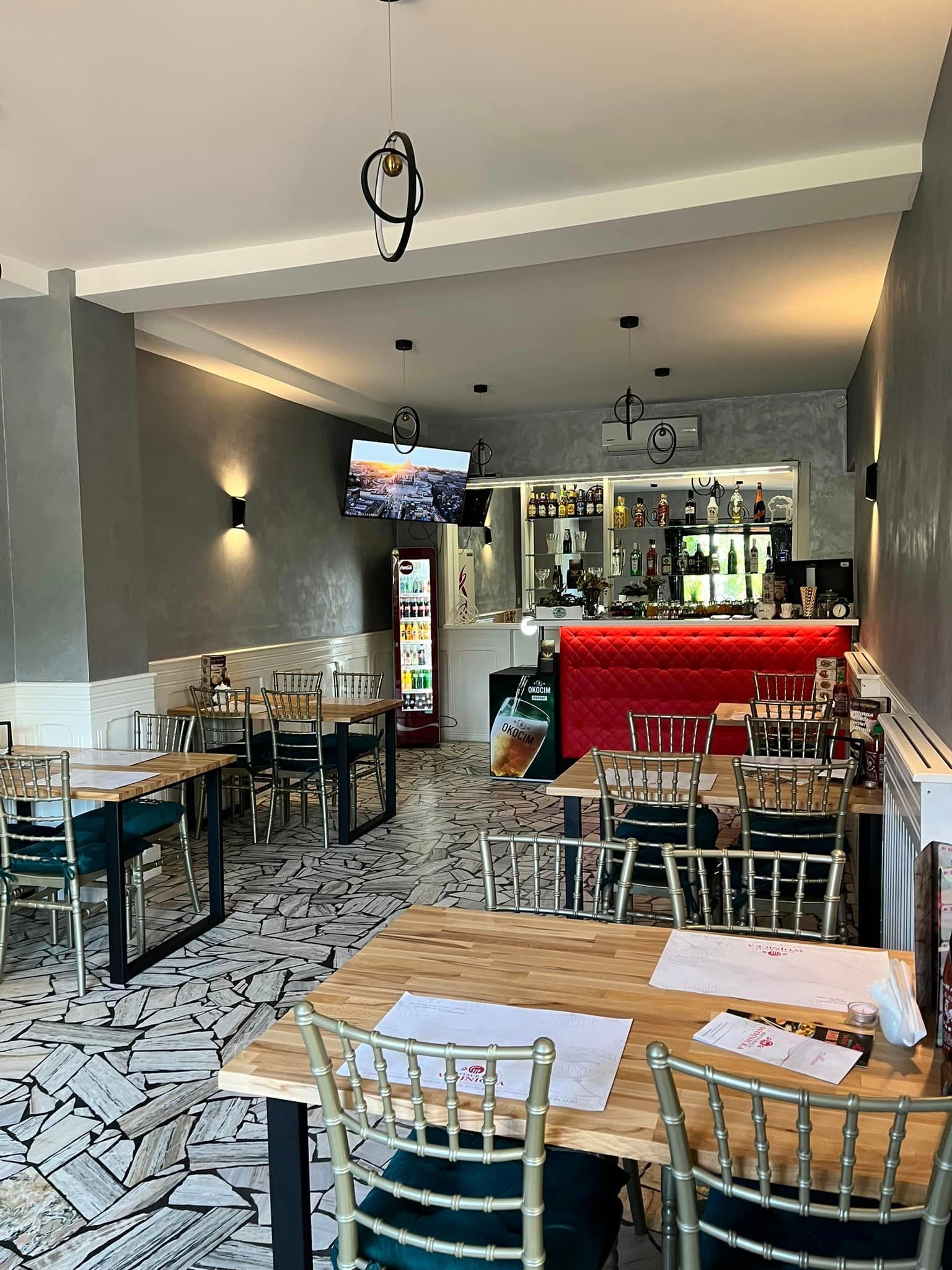 Wnętrze sali restauracyjnej z drewnianymi stołami nakrytymi menu, złotymi krzesłami z zielonym obiciem, barem z czerwonym podświetleniem w tle oraz dekoracyjnymi lampami sufitowymi.