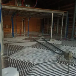 Montaż ogrzewania podłogowego na folii aluminiowej z widocznymi rurami grzewczymi i elementami konstrukcji budynku w tle.