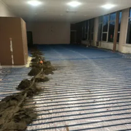 Montaż ogrzewania podłogowego w dużym pomieszczeniu, widoczne niebieskie rurki na folii z siatką oraz przygotowana zaprawa wzdłuż ściany.