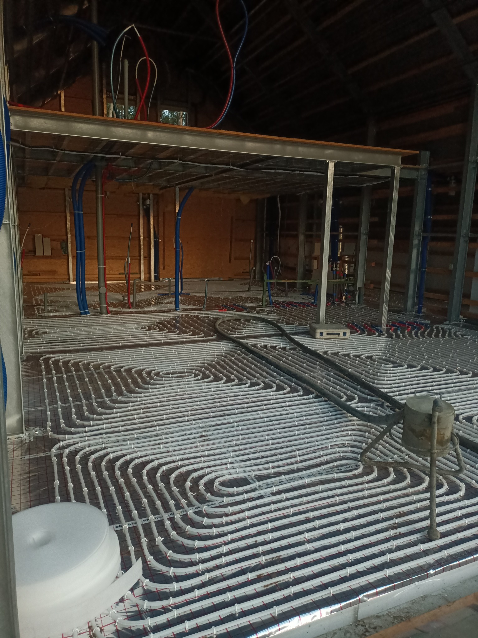 Montaż ogrzewania podłogowego na folii aluminiowej z widocznymi rurami grzewczymi i elementami konstrukcji budynku w tle.