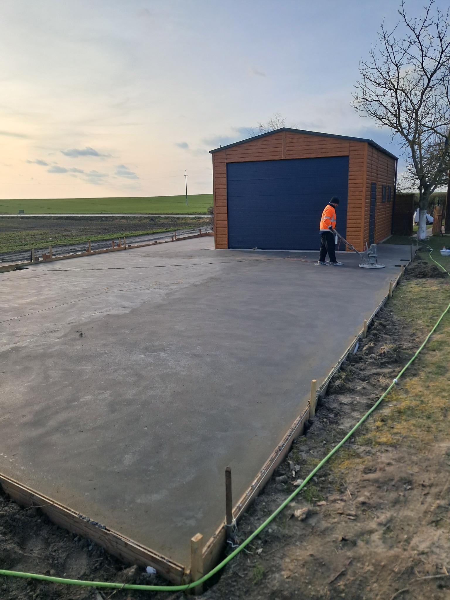 Pracownik w pomarańczowej kamizelce wyrównuje świeżo wylaną betonową posadzkę na zewnątrz budynku gospodarczego z garażem.