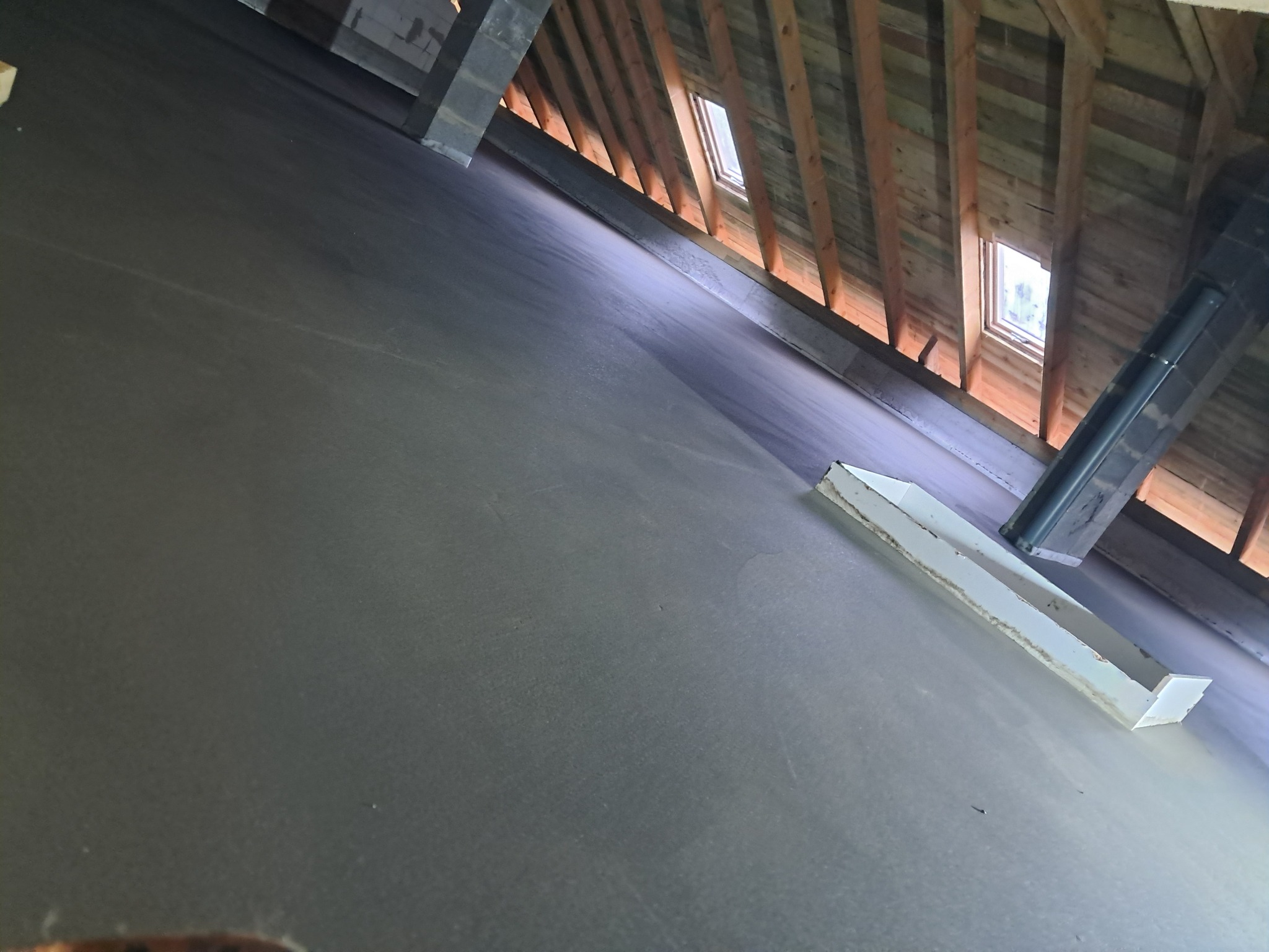 Gładka, szara powierzchnia wylanego jastrychu w poddaszu z drewnianą konstrukcją dachu i oknami połaciowymi. Widoczne elementy konstrukcyjne i narzędzia.