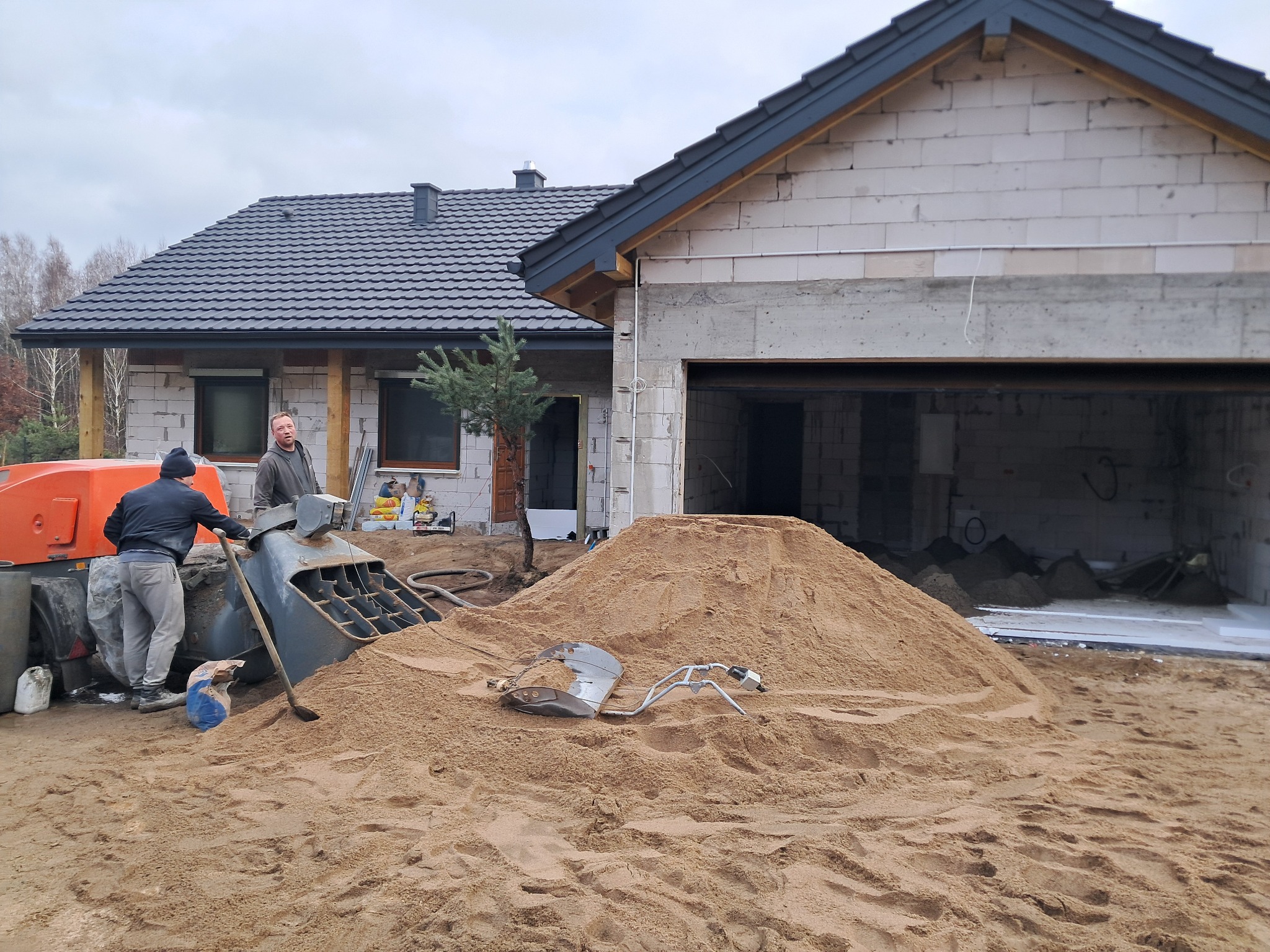 Prace przygotowawcze do wylewki jastrychowej: ekipa przy maszynie, góra piasku na placu budowy domu w stanie surowym otwartym, widoczne narzędzia.