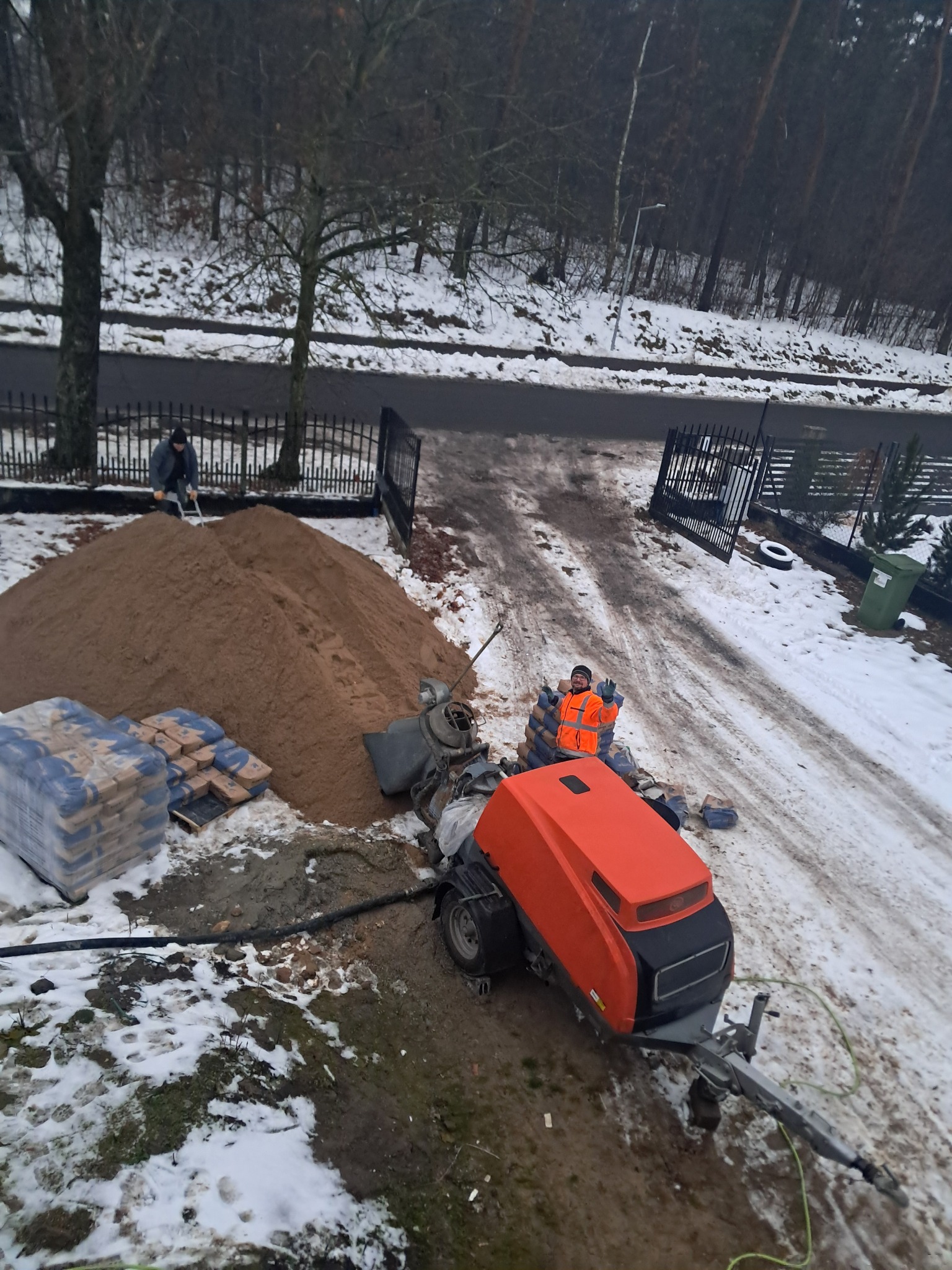 Prace przygotowawcze do wylewki: pomarańczowa maszyna do jastrychu, sterta piasku, worki z cementem na ośnieżonym terenie posesji. Pracownik w odblaskowej kamizelce.