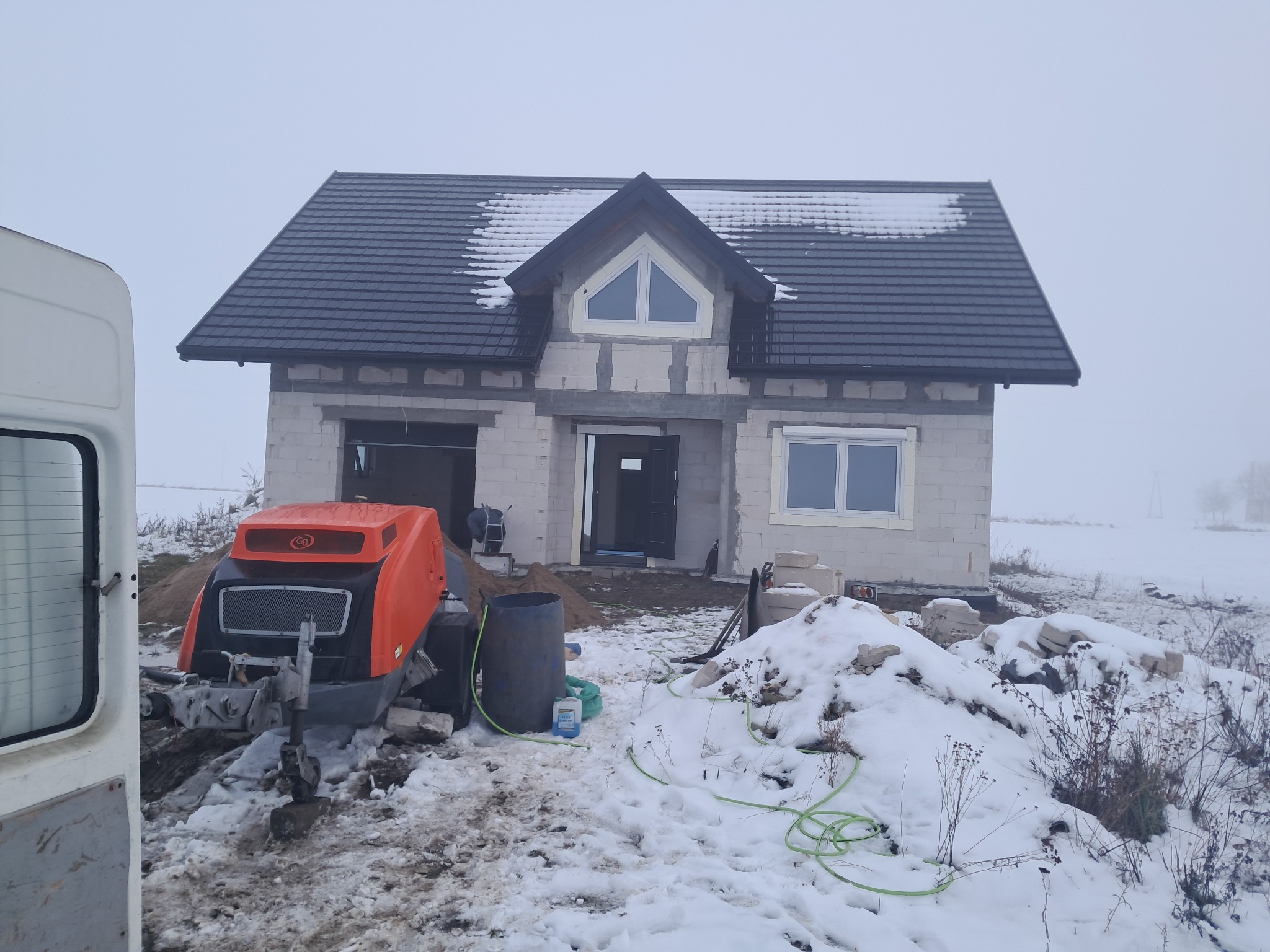 Budowa domu jednorodzinnego w zimowej scenerii. Widoczna maszyna do wylewania posadzek, ściany z betonu komórkowego i czarny dach z połaciami pokrytymi śniegiem.