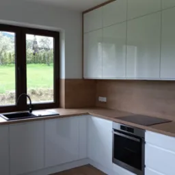 Nowoczesna kuchnia z białymi, lakierowanymi frontami szafek i drewnianym blatem, czarny zlew i bateria przy oknie z widokiem na zielony ogr&oacute;d.