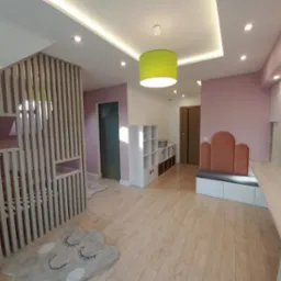 Pokój dziecięcy z antresolą zabezpieczoną drewnianą konstrukcją z pionowych listew, doświetlony zieloną lampą wiszącą, z dywanem w kształcie chmury i zabudową meblową pod ścianą.