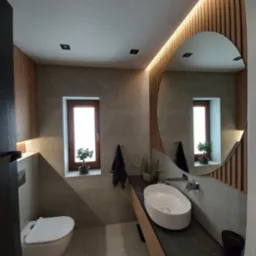 Nowoczesna łazienka z owalną umywalką na blacie z ciemnego kamienia, okrągłe lustro z podświetleniem LED, drewniane lamele na ścianie i wisząca toaleta.