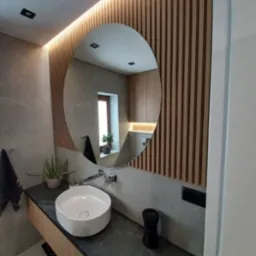 Nowoczesna łazienka z okrągłym lustrem, drewnianymi lamelami na ścianie i umywalką nablatową. W lustrze odbija się okno z rośliną. Blat z ciemnego kamienia.