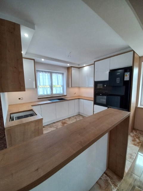 Nowoczesna kuchnia z białymi błyszczącymi szafkami i drewnianymi blatami, widoczny fragment wyspy kuchennej z blatem w kolorze jasnego drewna, czarny sprzęt AGD w zabudowie.