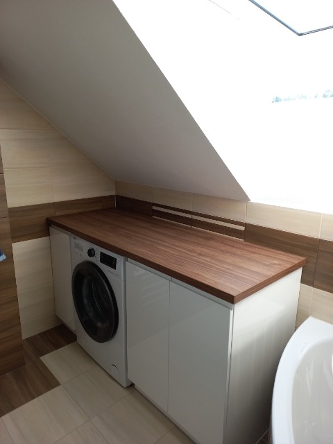 Aranżacja pralki zabudowanej w szafce pod blatem drewnianym, w łazience ze skośnym sufitem w Laskowej.