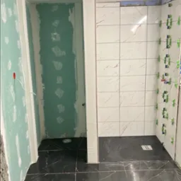 Łazienka w trakcie wykończenia: czarne płytki podłogowe z białym żyłowaniem, białe płytki ścienne imitujące marmur, zielone krzyżyki glazurnicze, surowe ściany przygotowane pod malowanie.