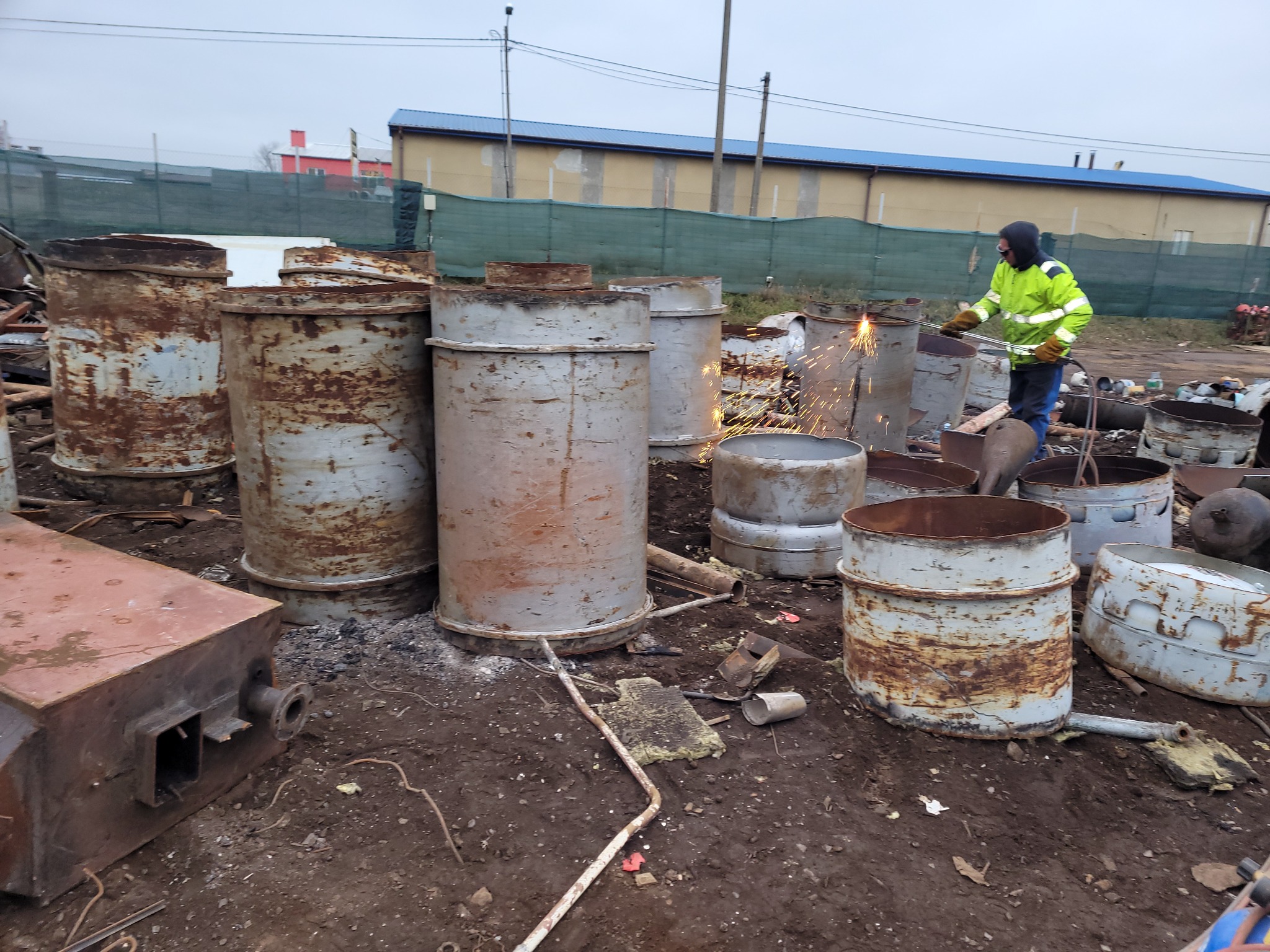 Pracownik w odblaskowej kurtce tnący metalową beczkę palnikiem w miejscu składowania złomu, widoczne iskry i rdzawe beczki.