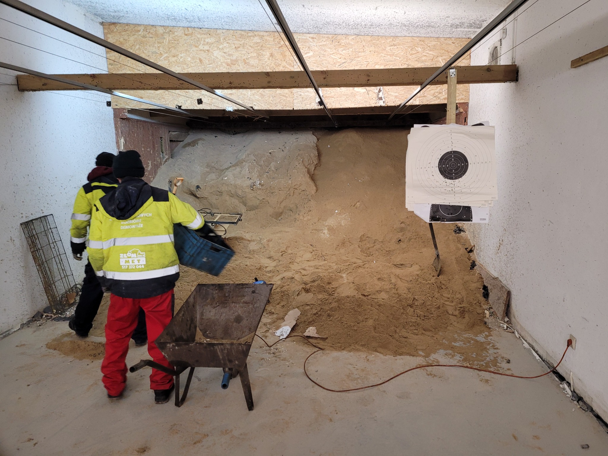 Wnętrze pomieszczenia z dużą ilością piasku, dwoma pracownikami w odblaskowych kurtkach i czerwoną taczką, widoczne tarcze strzelnicze na ścianie.