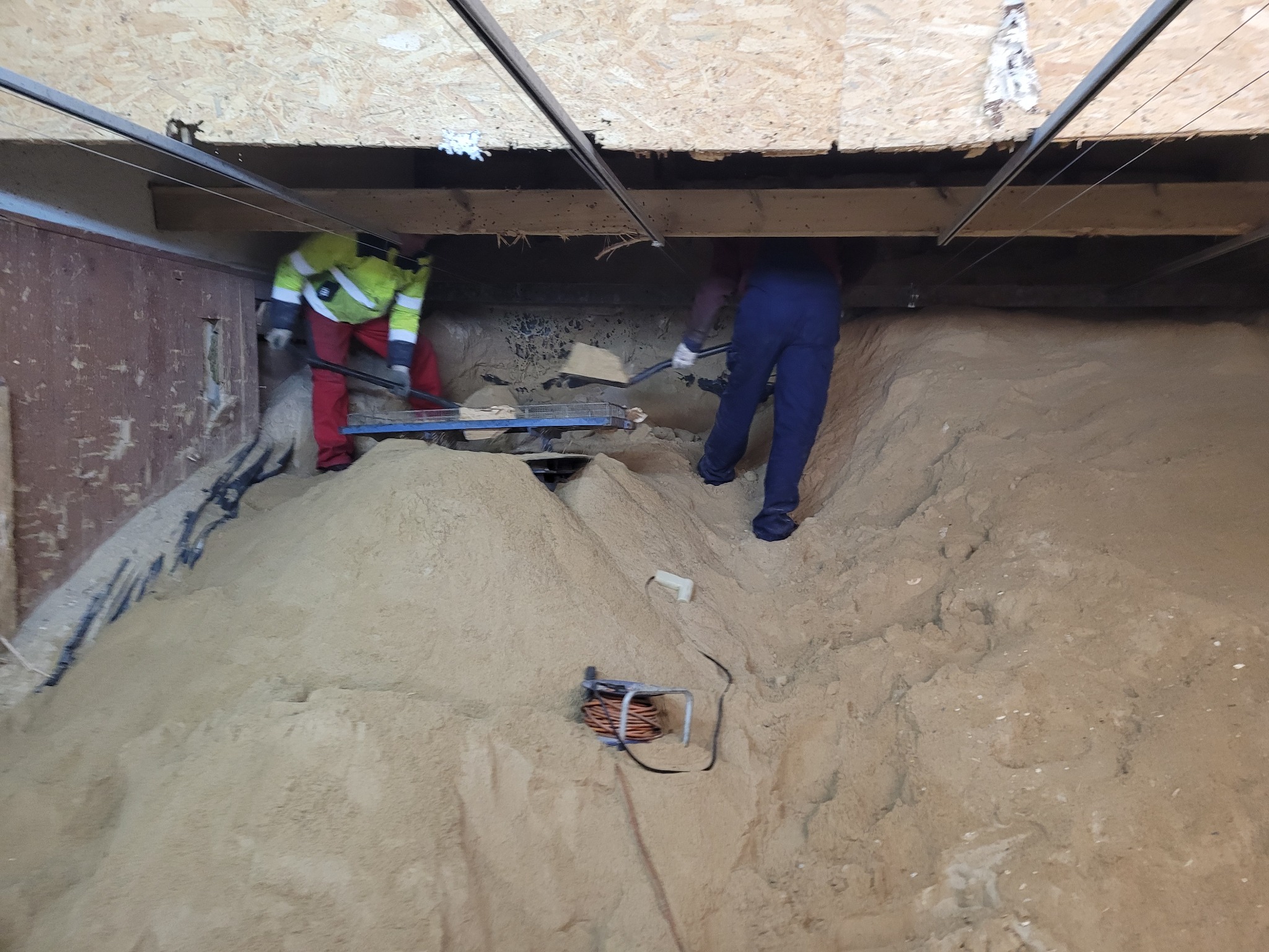 Dwóch pracowników usuwa piasek z przestrzeni pod podłogą, widoczne narzędzia i zwój kabla elektrycznego na piasku.
