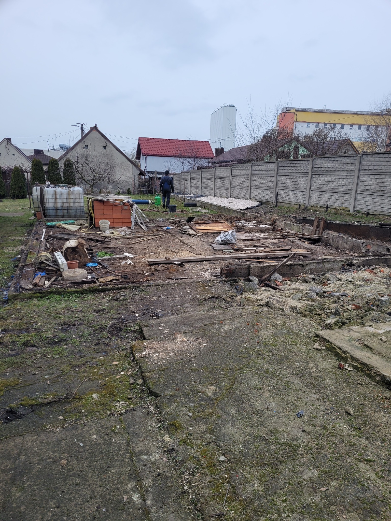 Teren po rozbiórce budynku z widocznymi fundamentami, gruzem, stertą cegieł i zbiornikiem na wodę w tle, osoba oddalająca się w kierunku ogrodzenia.