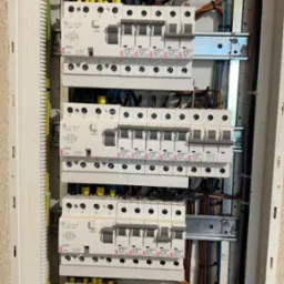 Rozdzielnica elektryczna z zainstalowanymi bezpiecznikami i okablowaniem wewnątrz białej obudowy, widoczne różne kolory przewodów i elementy mocujące.