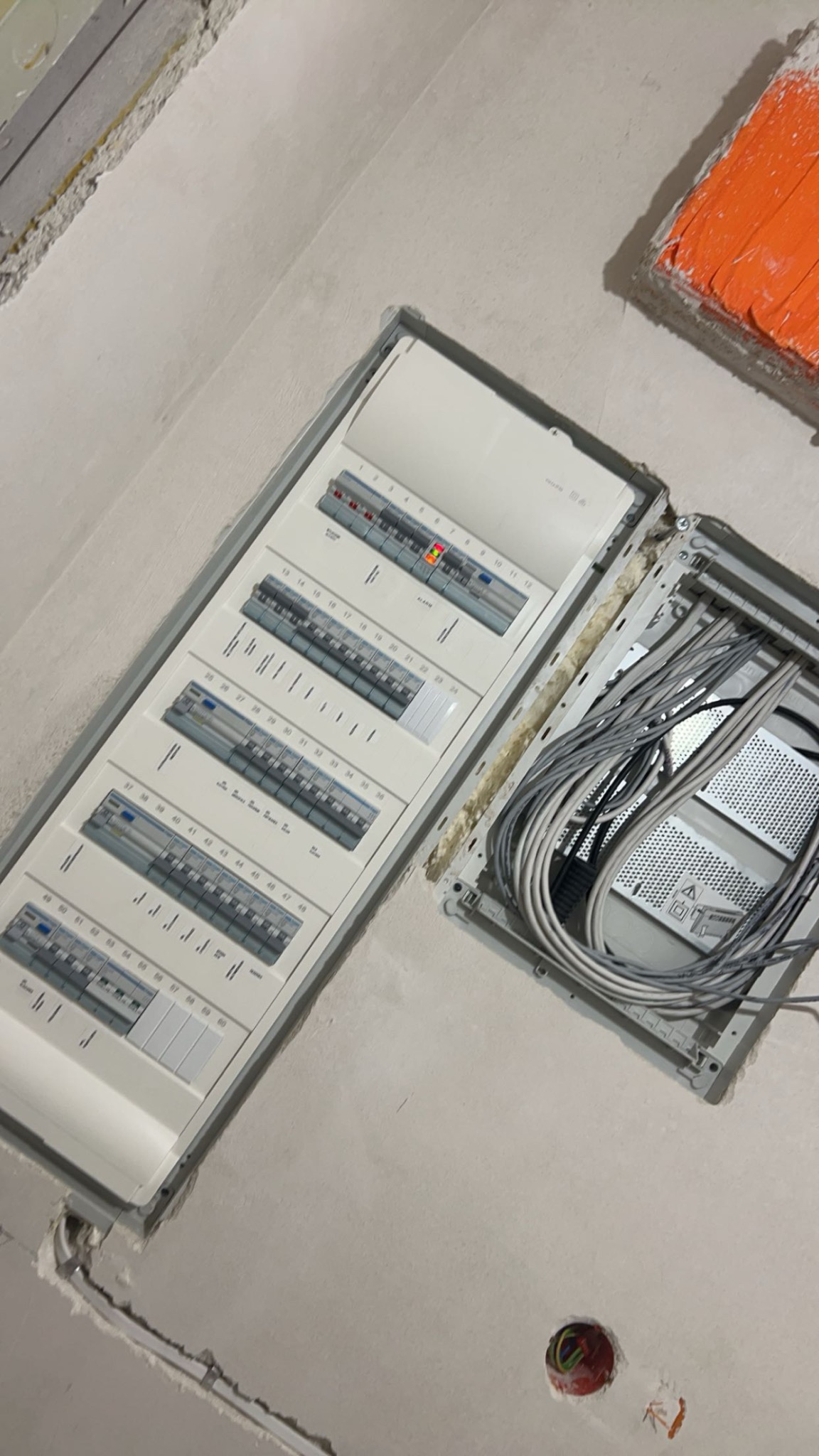 Rozdzielnica elektryczna z otwartą obudową, widoczne przewody wewnątrz, zamontowana w surowej ścianie podczas prac wykończeniowych.