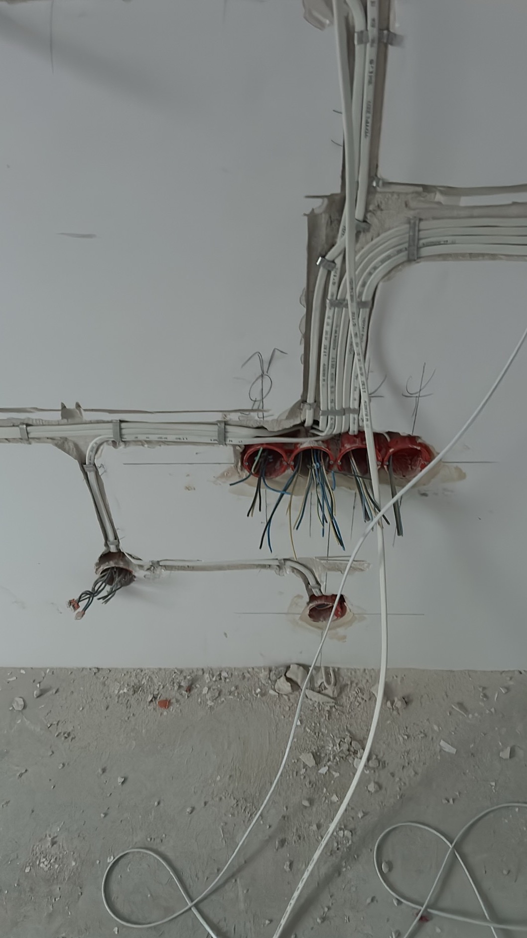 Rozprowadzone kable elektryczne w ścianie i suficie, widoczne puszki instalacyjne z wystającymi przewodami, ślady po kuciu w ścianie, instalacja w trakcie realizacji.
