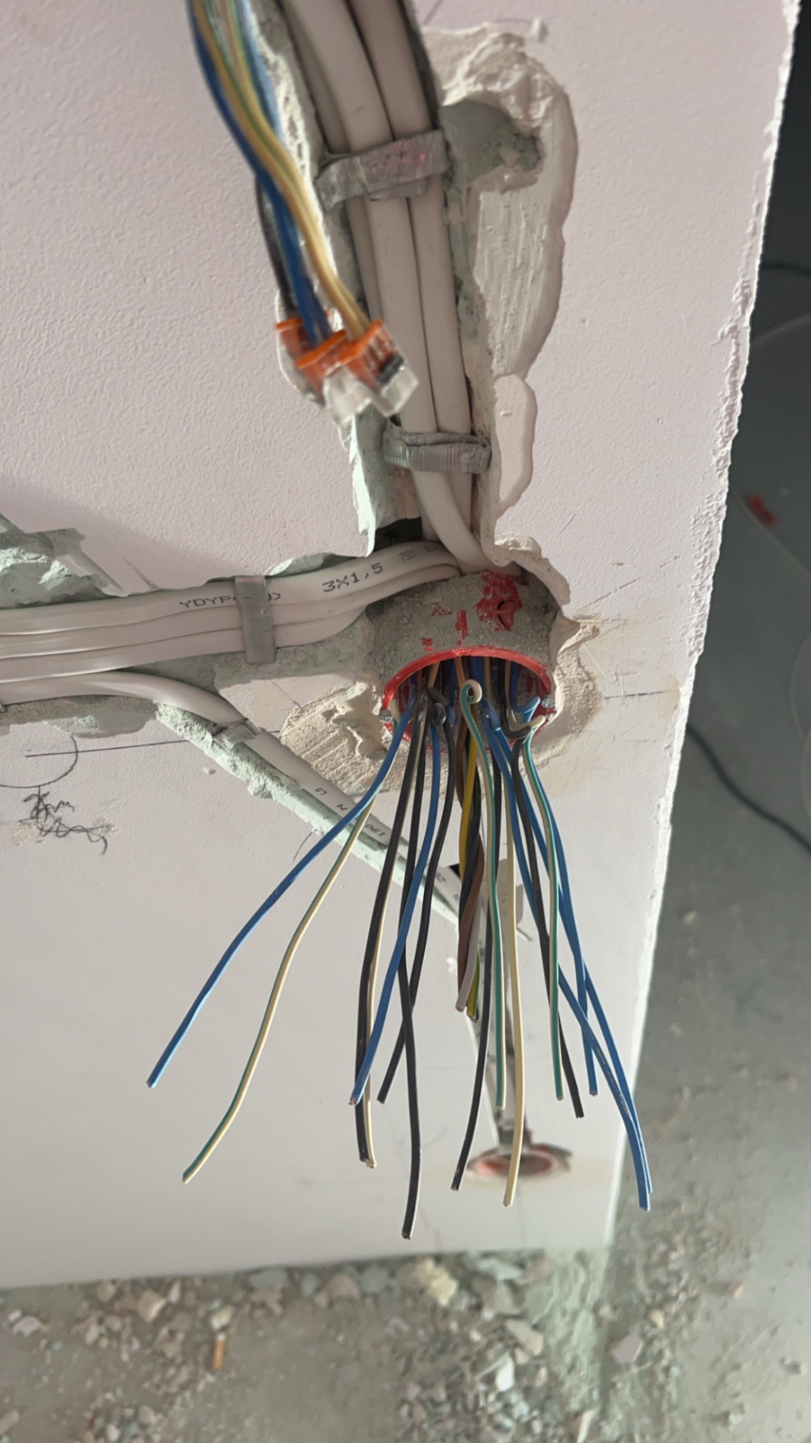 Wiązka kolorowych przewodów wystająca z okrągłego otworu w suficie, widoczne kable prowadzone w listwach mocowanych do sufitu, surowy stan wykończenia.