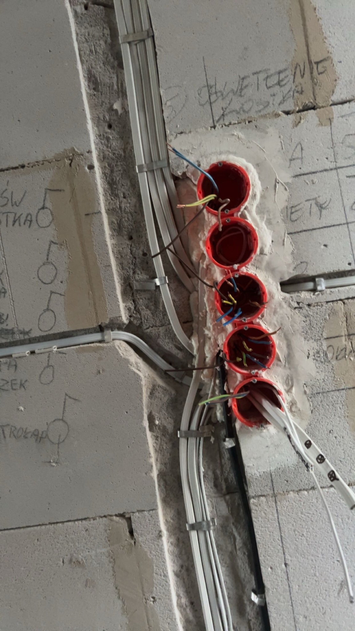 Pionowy układ czterech czerwonych puszek instalacyjnych w surowej ścianie, widoczne przewody elektryczne w różnych kolorach oraz białe kable przymocowane metalowymi uchwytami, na ścianie odręczne...