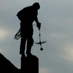 Sylwetka osoby w kombinezonie stojącej na kominie z narzędziami do czyszczenia na tle pochmurnego nieba.