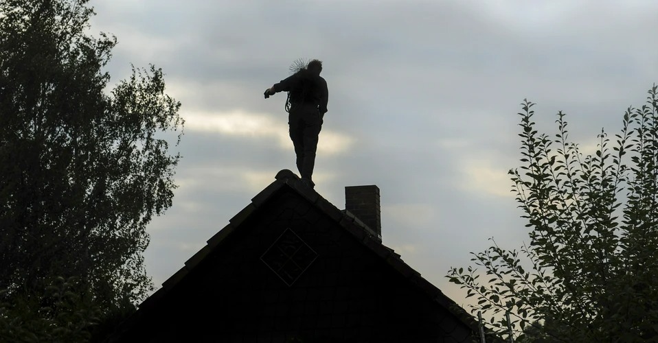 Sylwetka osoby na dachu domu z kominem, trzymającej narzędzie do czyszczenia komina, na tle pochmurnego nieba.