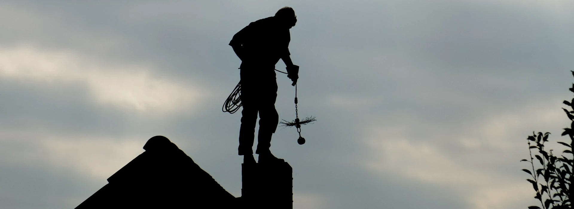 Sylwetka osoby w kombinezonie stojącej na kominie z narzędziami do czyszczenia na tle pochmurnego nieba.