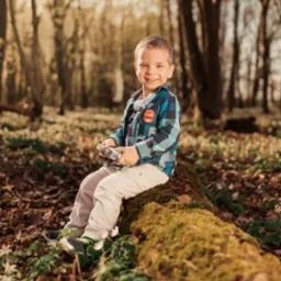 Uśmiechnięty chłopiec w kraciastej koszuli siedzi na omszałym pniu drzewa w lesie, trzymając stary aparat fotograficzny.