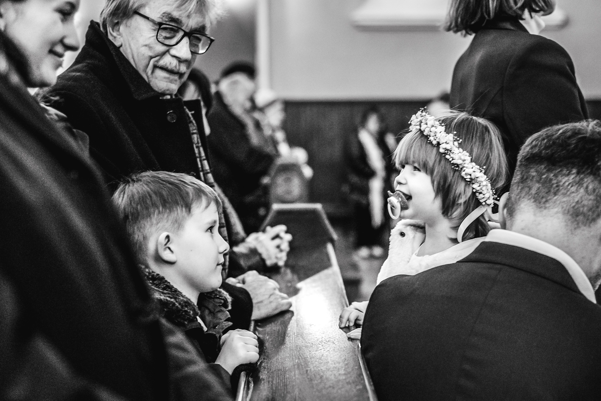 Czarno-białe zdjęcie z uroczystości chrztu: dziewczynka w wianku na głowie z smoczkiem w ustach, obok chłopiec zamyślony, a za nimi uśmiechnięty dziadek w okularach.
