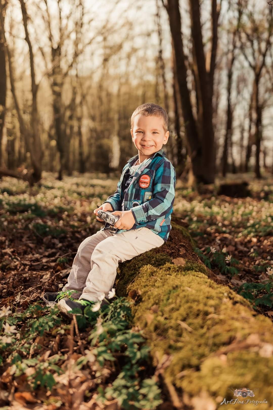 Uśmiechnięty chłopiec w kraciastej koszuli siedzi na omszałym pniu drzewa w lesie, trzymając stary aparat fotograficzny.